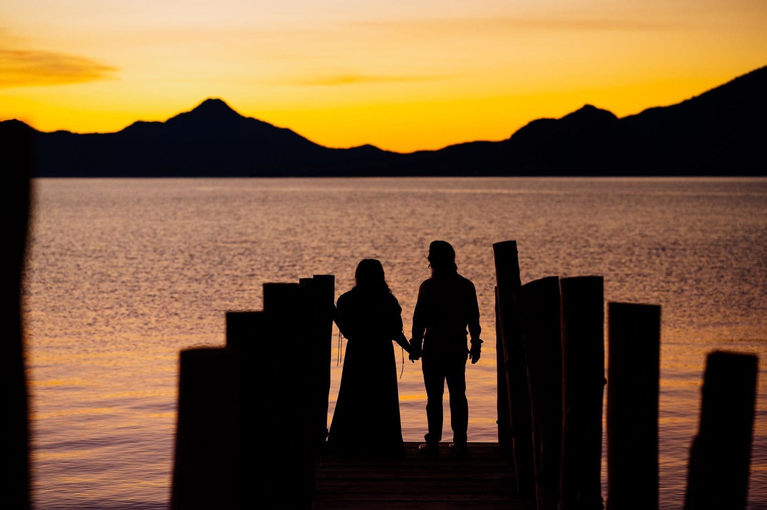 Couple silhouetted hand-in-hand on a wooden pier against an orange sunset sky over Lake Atitlan.