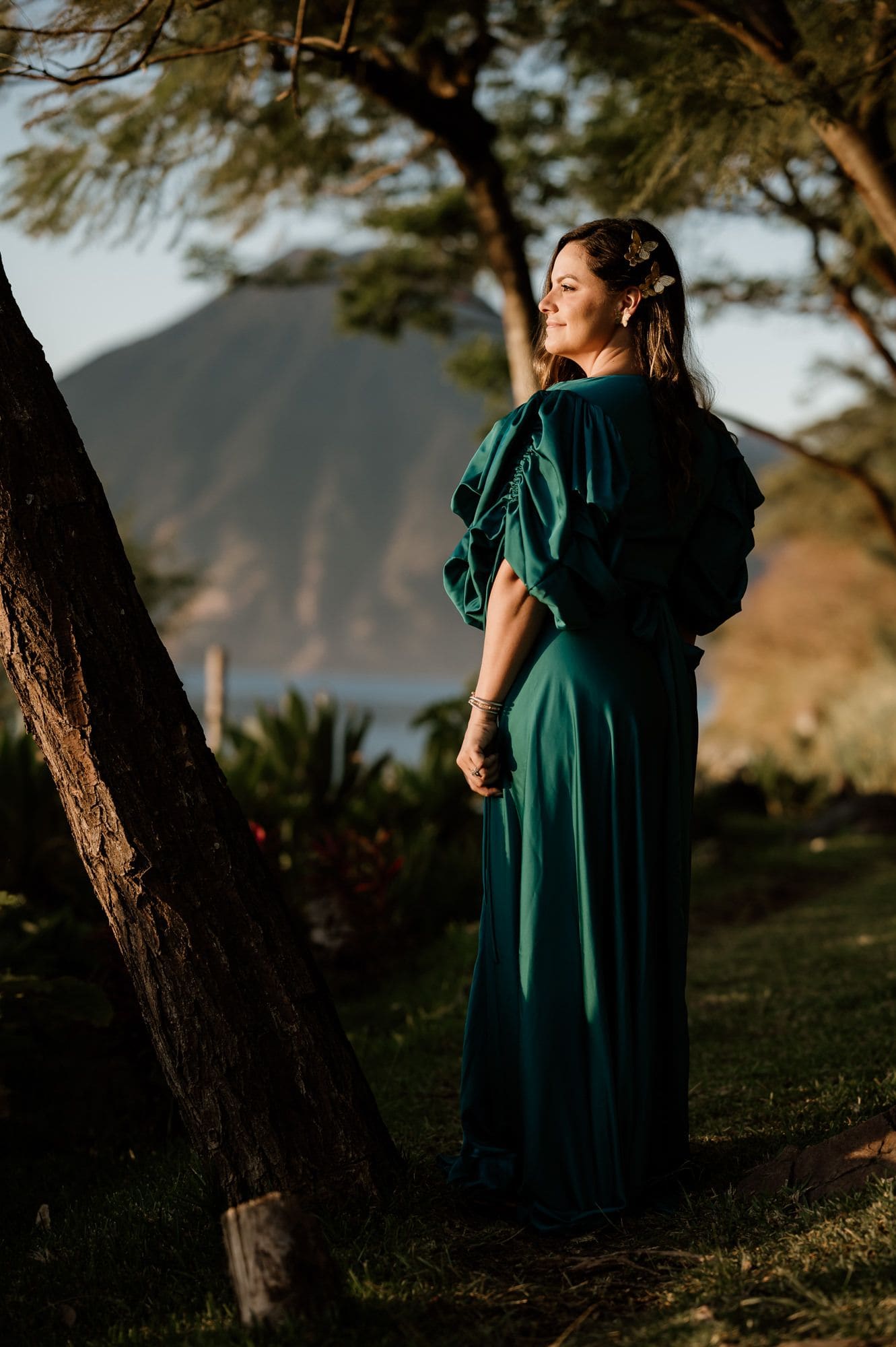 Bride glances back in a flowing emerald gown framed by trees with a volcano in the distance during the Lake Atitlán pre-wedding session.