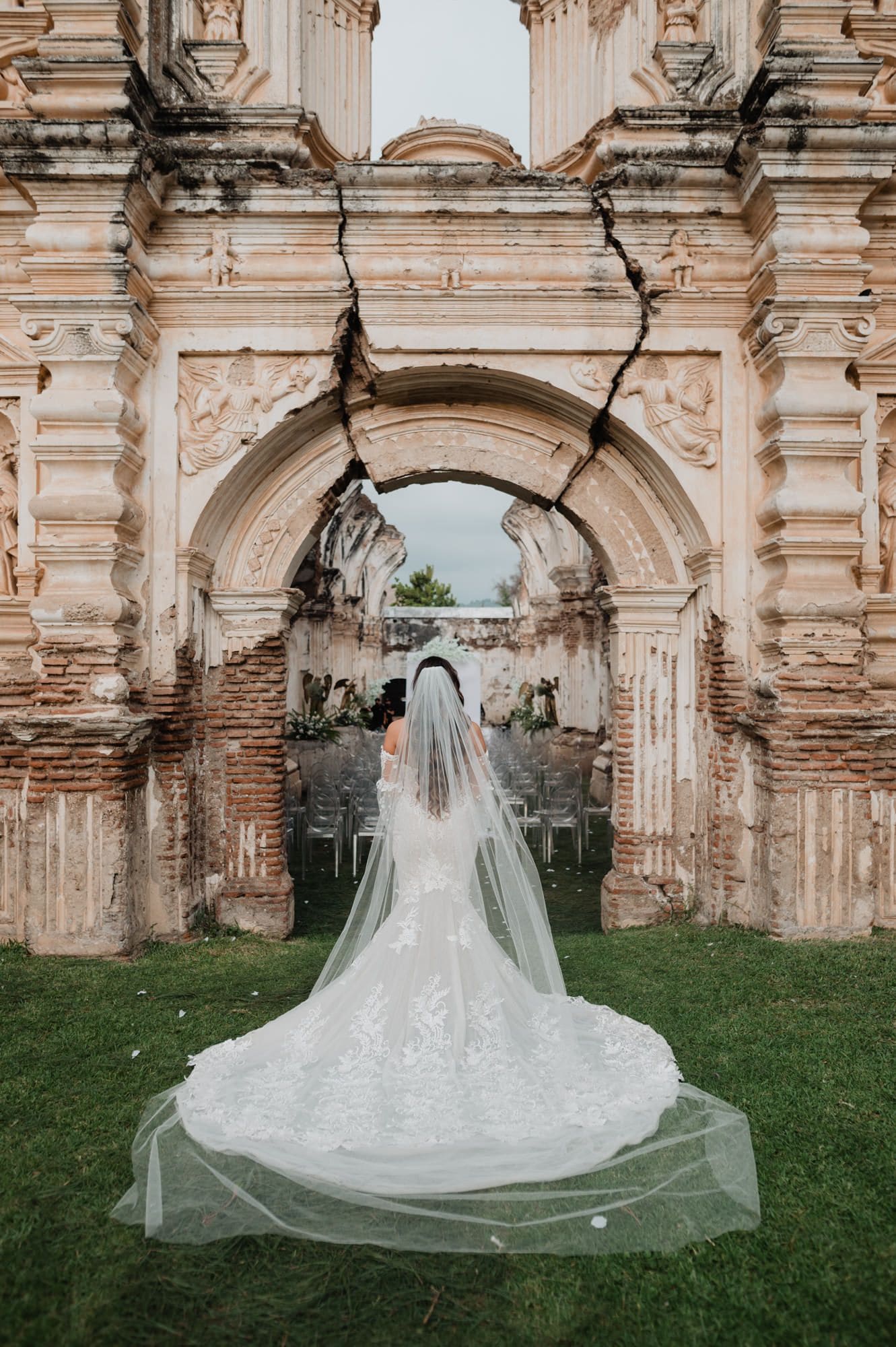 Bride walks away from the camera through a cracked baroque archway with her veil flowing onto the grass.
