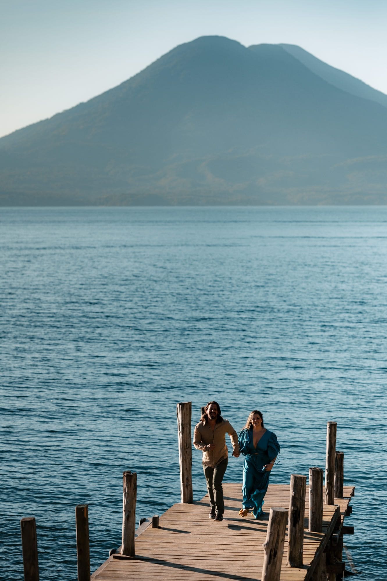 Couple holds hands running down a wooden dock toward the camera with the volcano rising behind Lake Atitlán during their pre-wedding session.