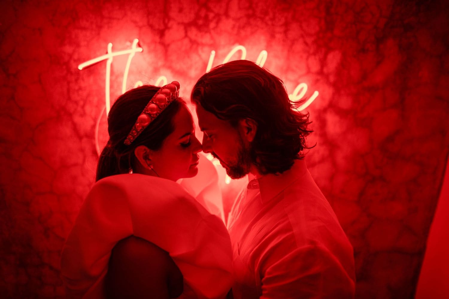 Bride and groom touch foreheads under a red neon sign casting cinematic crimson light over the photo booth wall at the reception.