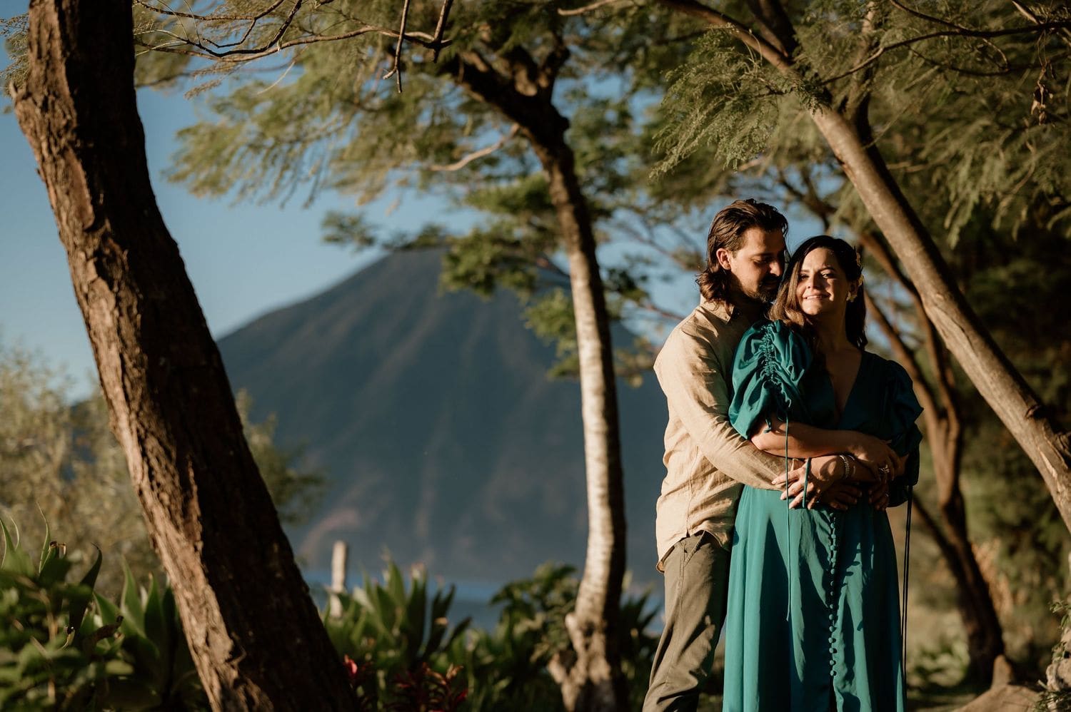 Groom hugs the bride from behind among acacia branches with the volcano rising in the background at Lake Atitlán.
