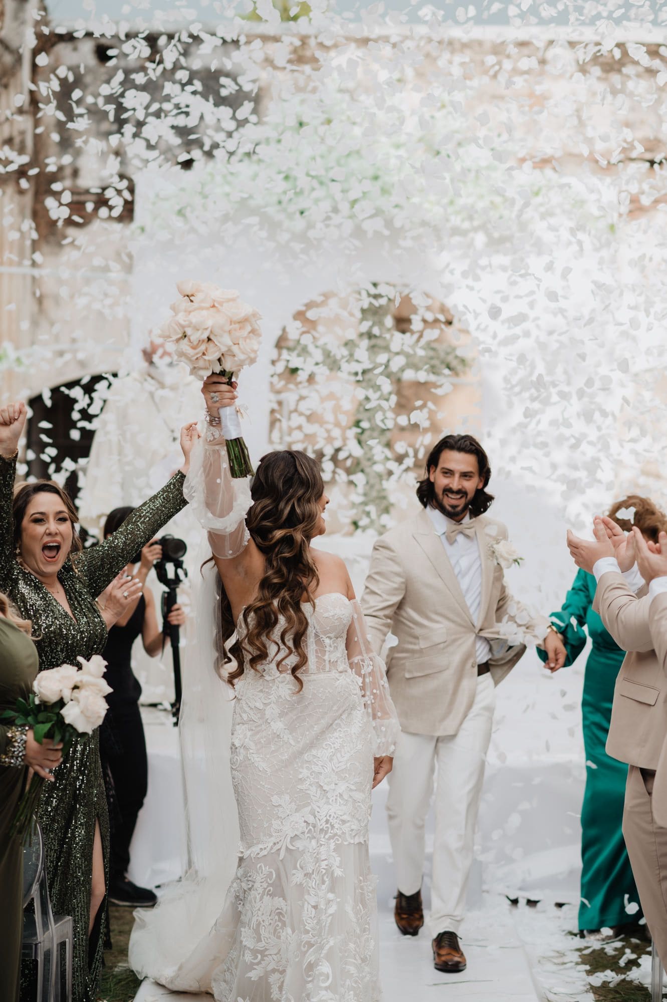 Bride raises her bouquet from behind as the couple recess down the aisle under a shower of white petal confetti at Casa de la Ruina.