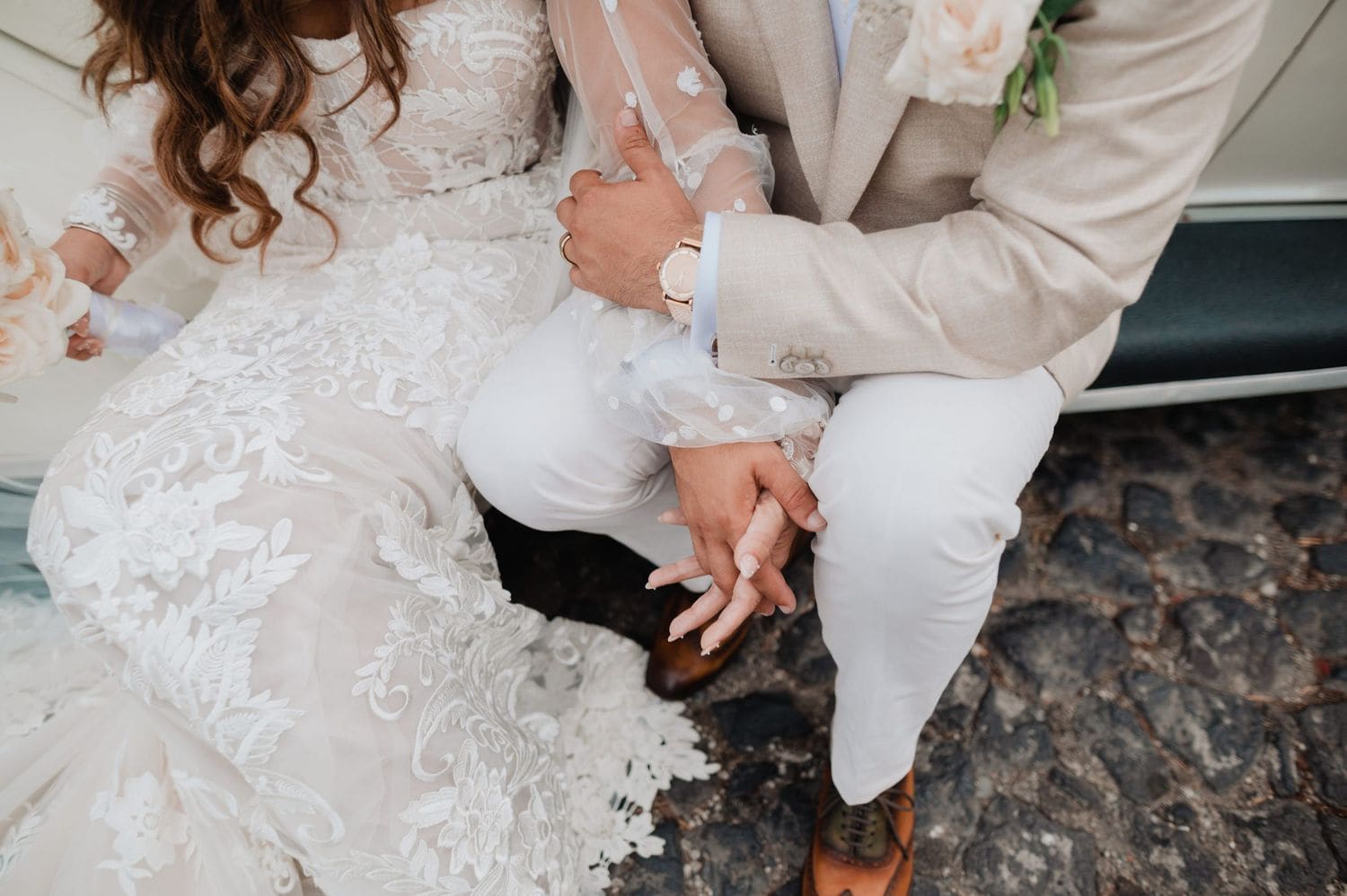 Detail of joined hands resting on the couple's laps showing the wedding band, watch and lace cuff against the cobblestones.