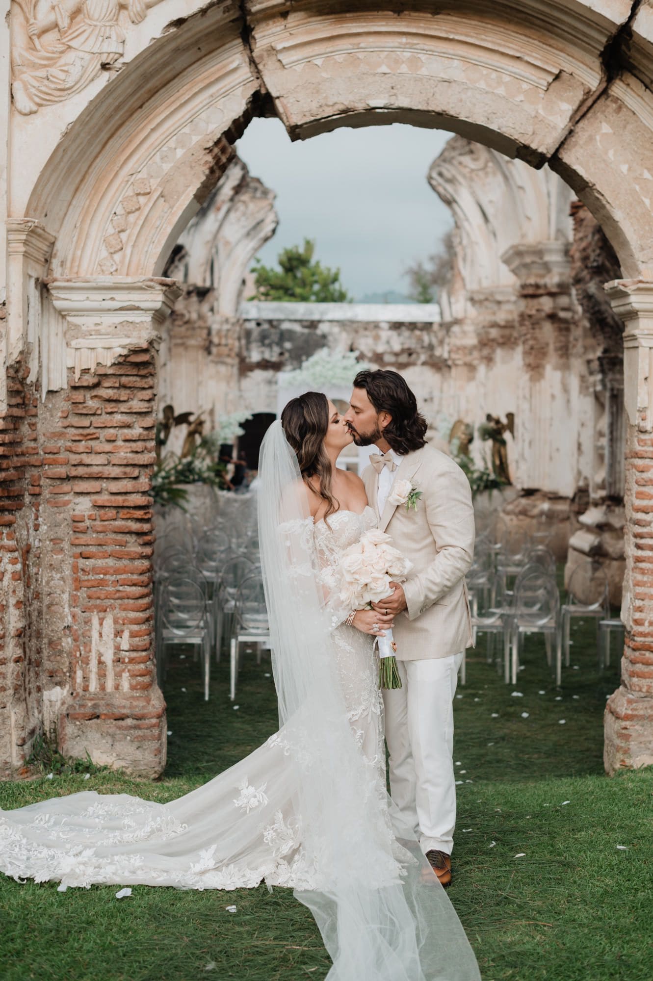 Bride and groom share a first kiss framed by a baroque ruin arch with chair rows and petals at Casa de la Ruina, Antigua Guatemala.