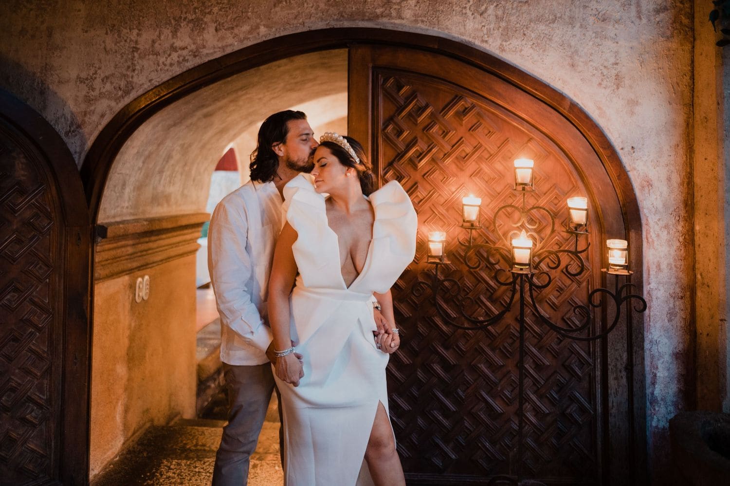 Bride in sculptural dress shares an intimate moment with the groom beside an arched stone passage lit by wrought-iron candle sconces.
