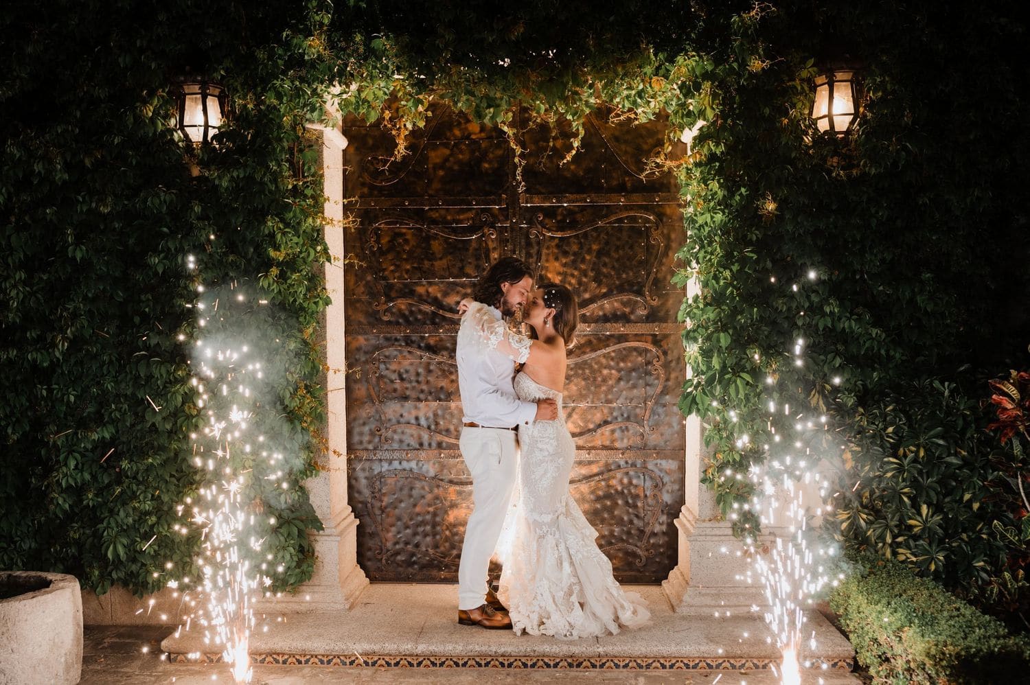 Bride and groom share a kiss between two columns of sparkling cold fountains under an ivy-covered arch at Hotel Pensativo.