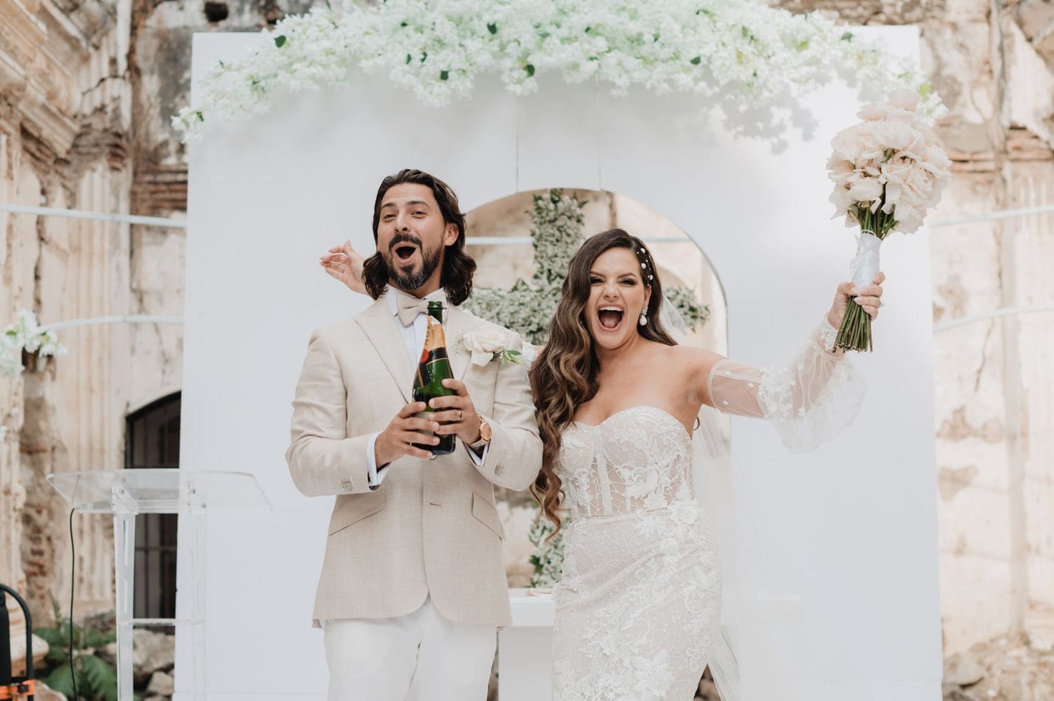 Groom pops champagne while the bride raises her bouquet and cheers at the white floral altar in Antigua Guatemala.