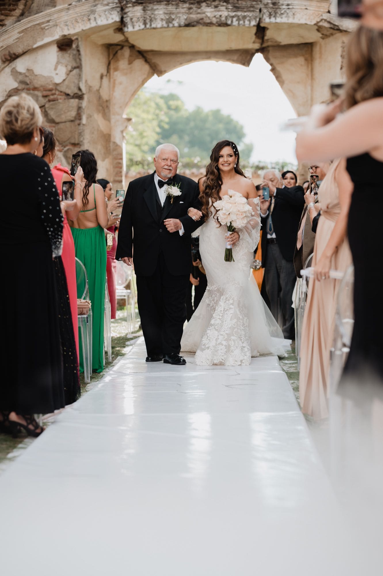Bride Marite walks down the aisle on her father's arm carrying a white bouquet under the ruin arch at Casa de la Ruina, Antigua Guatemala.
