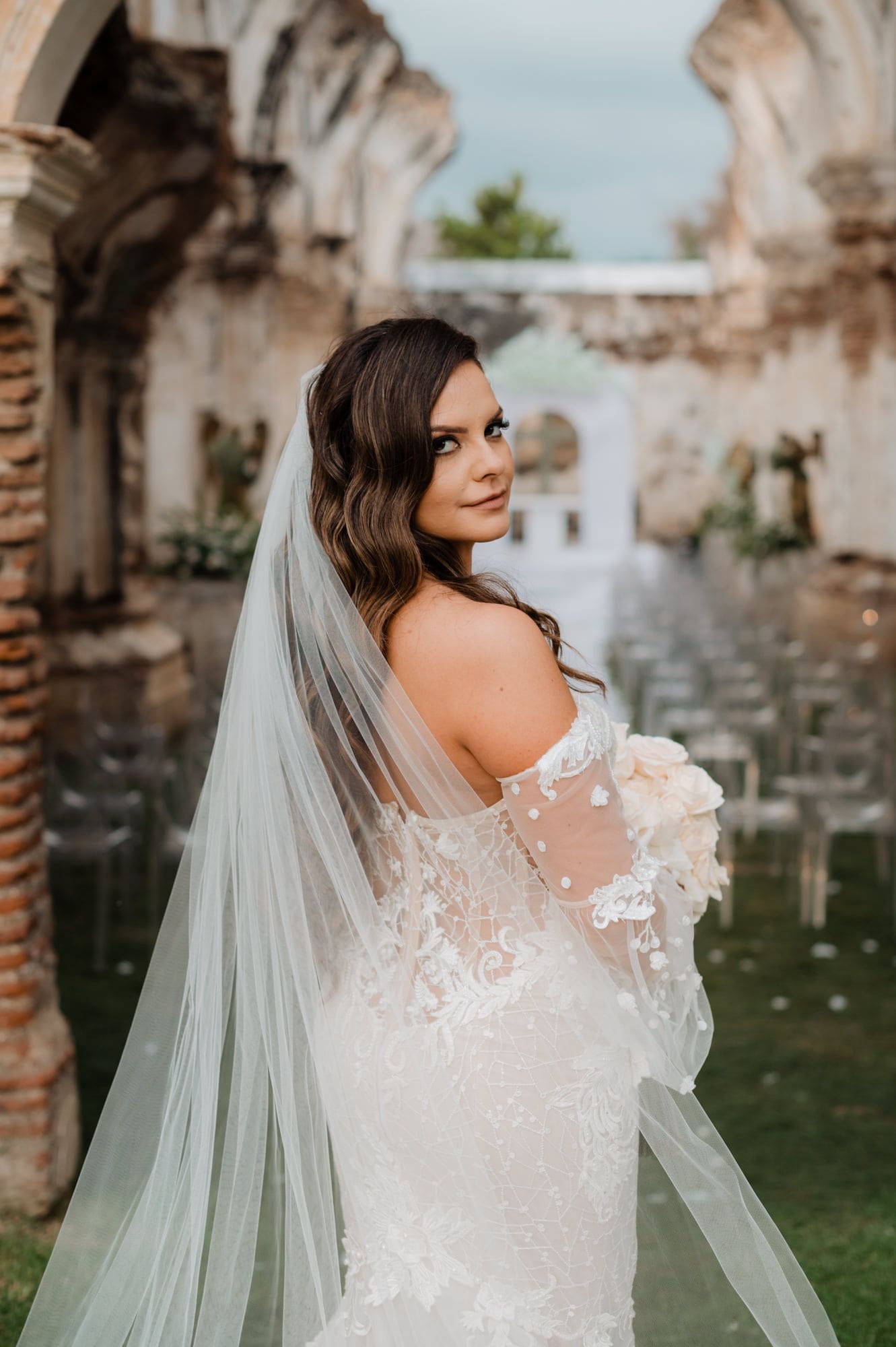 Marite glances back over her shoulder showing the off-shoulder lace bodice and trailing veil among colonial ruin arches.