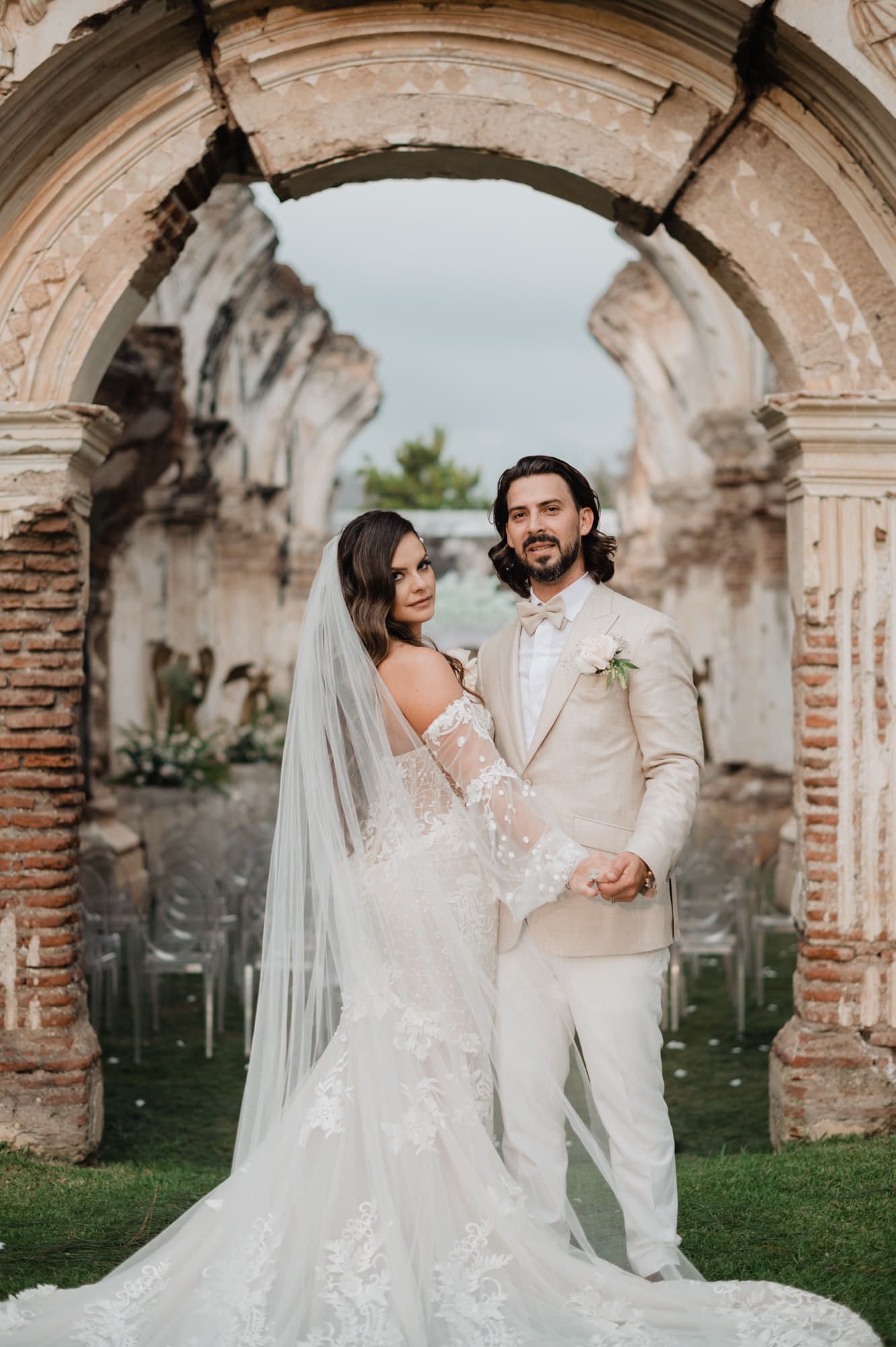 Couple poses confidently together centered under a tall brick ruin arch.