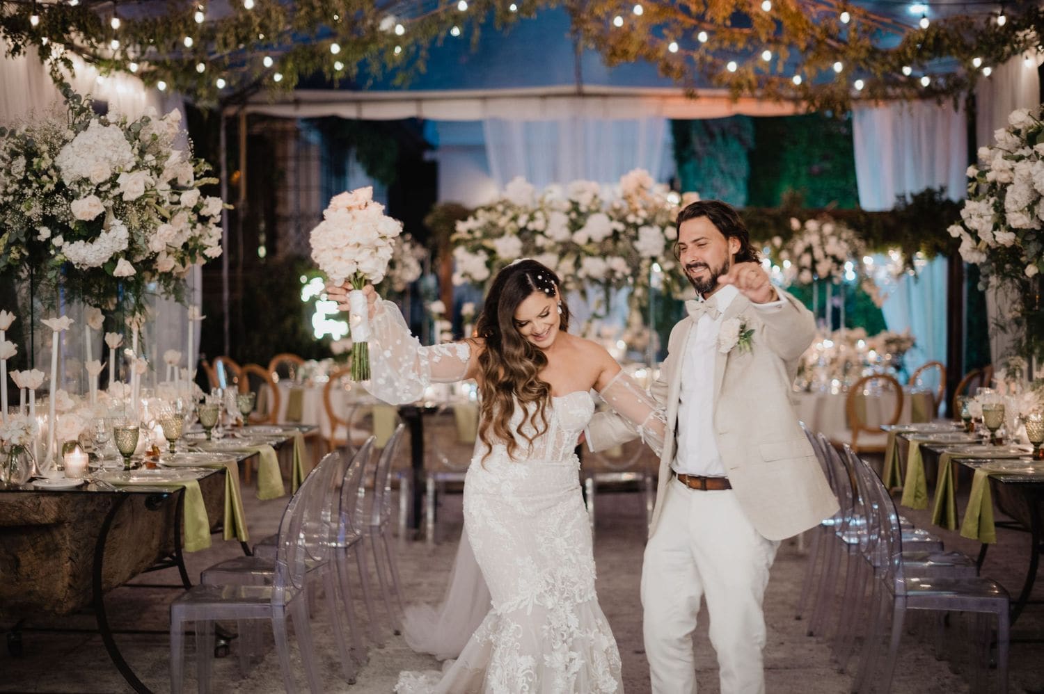 Bride and groom make their grand entrance between long banquet tables under hanging fairy lights and greenery at Hotel Pensativo.