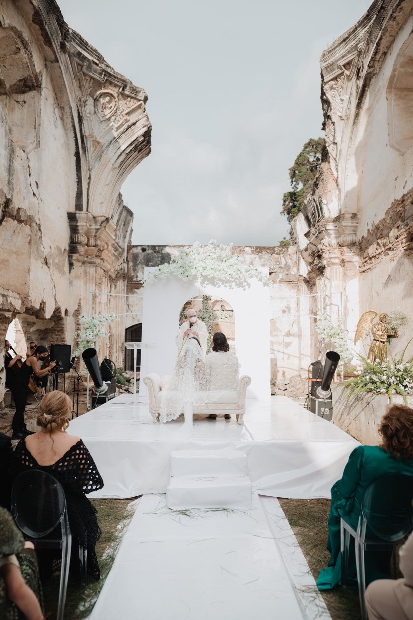 Wide view from the back of the aisle showing the couple at the altar between baroque ruined arches during the ceremony at Casa de la Ruina.