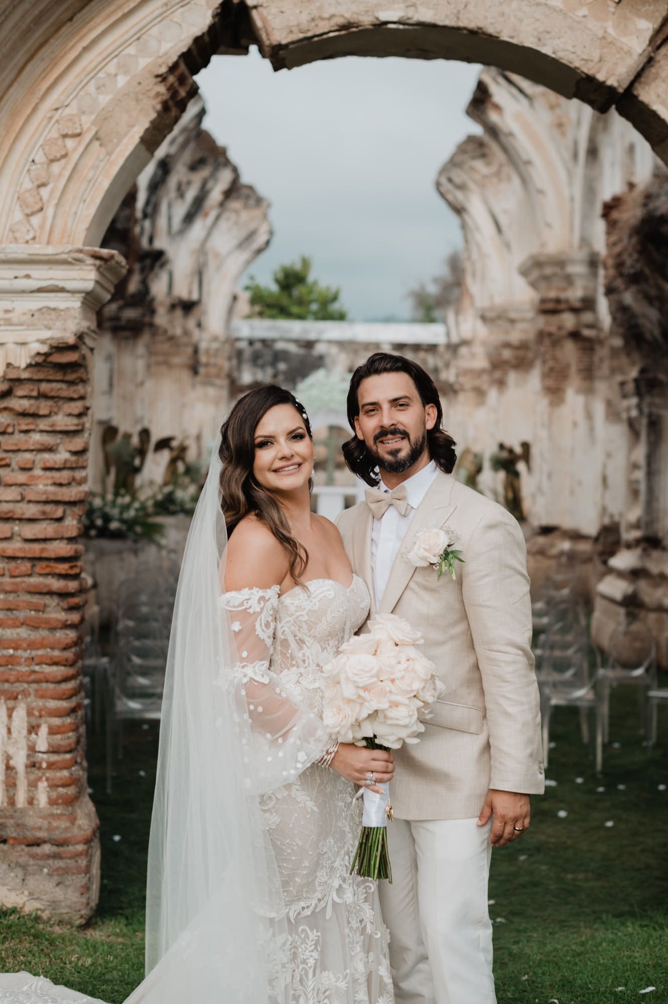 Editorial couple portrait under a baroque ruin arch with the train of the lace gown spread across the grass at Casa de la Ruina.