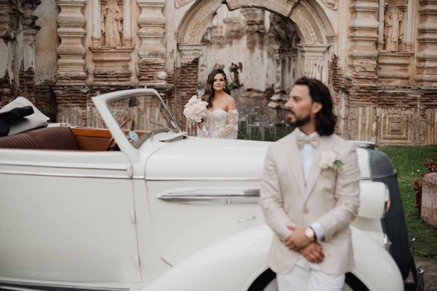 Bride stands inside a vintage white convertible holding her bouquet while the groom leans on the bumper in the foreground.