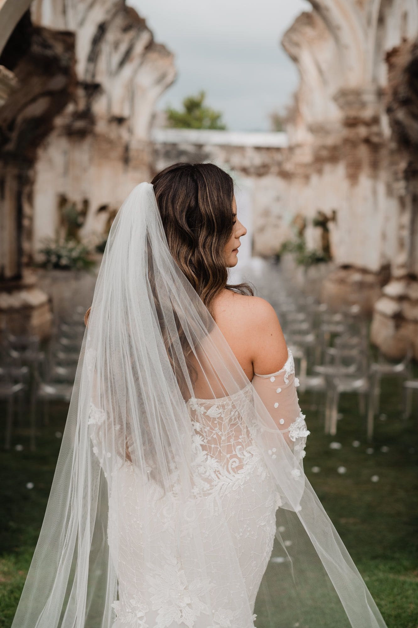 Back view of the bride showing her cathedral veil, off-shoulder lace sleeves and bare shoulders inside the ruin nave.