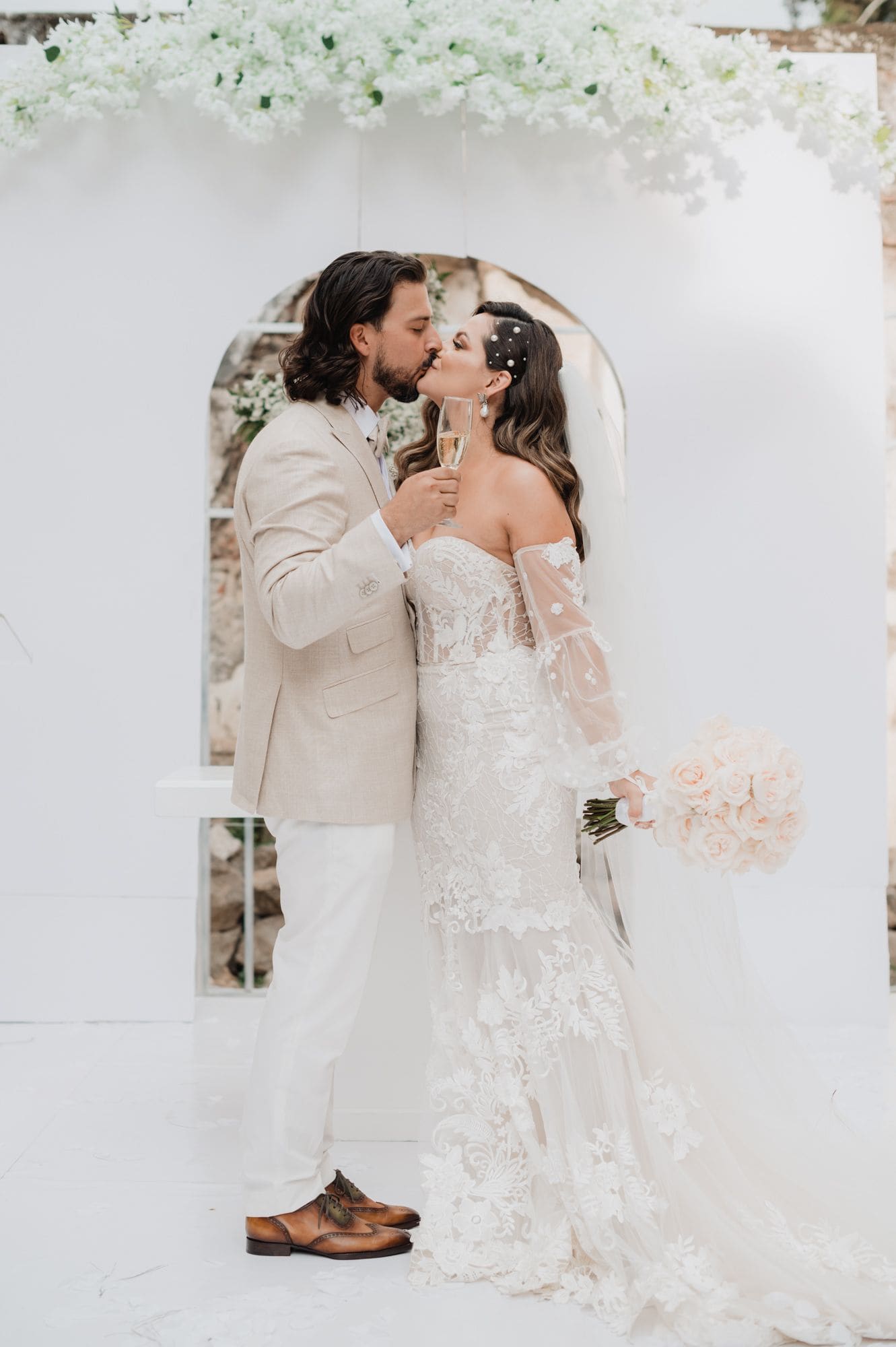 Couple toasts and kisses under the white flower arch with a glass of champagne and a blush rose bouquet after the ceremony.