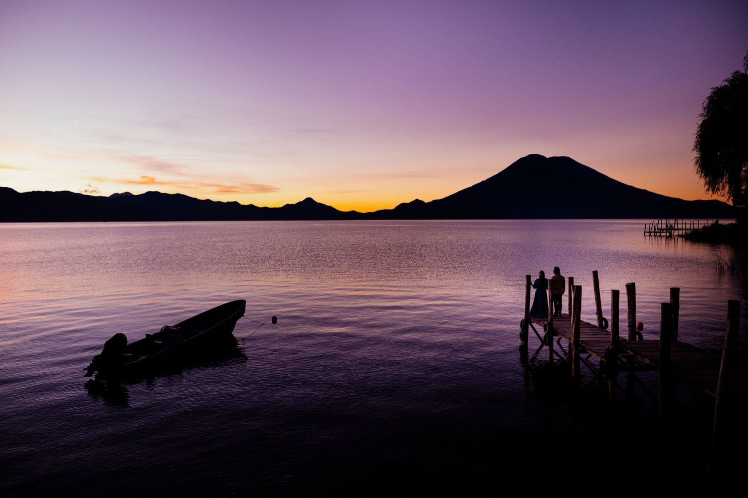 Silhouette of the couple at the end of a wooden pier with the volcano reflected on the violet sunset waters of Lake Atitlan.