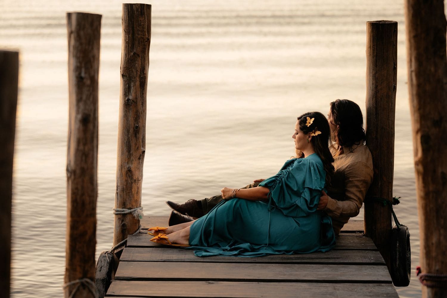 Bride in teal dress reclines against the groom on a wooden dock as warm light glows over Lake Atitlan.