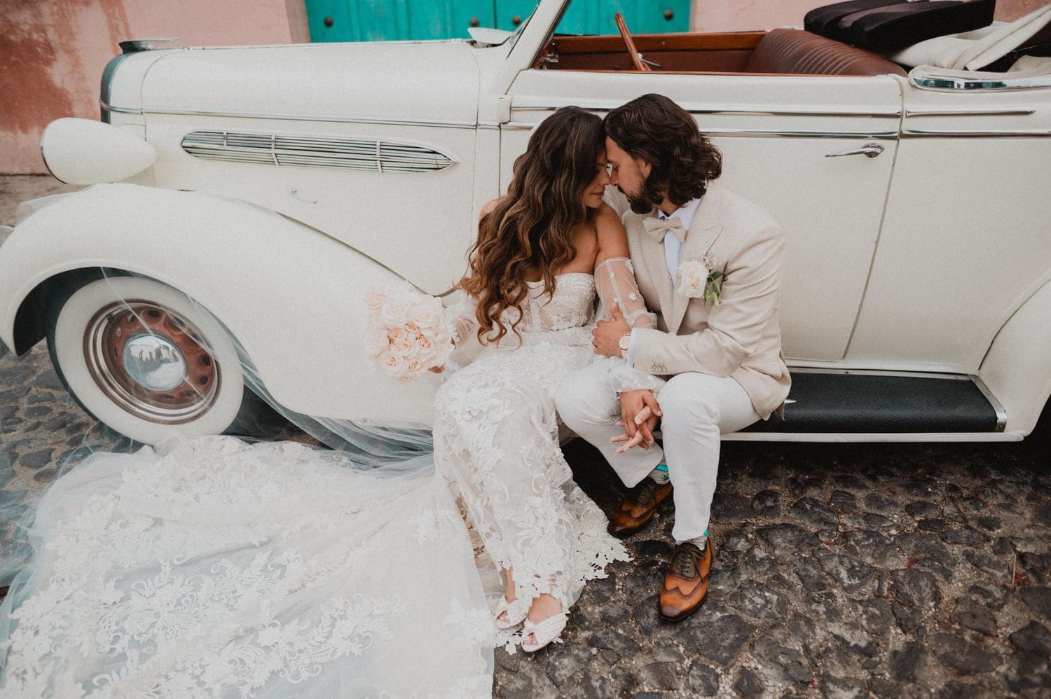 Couple sits intimately on the running board of a vintage car on Antigua's cobblestone street with the lace train spreading across the stones.