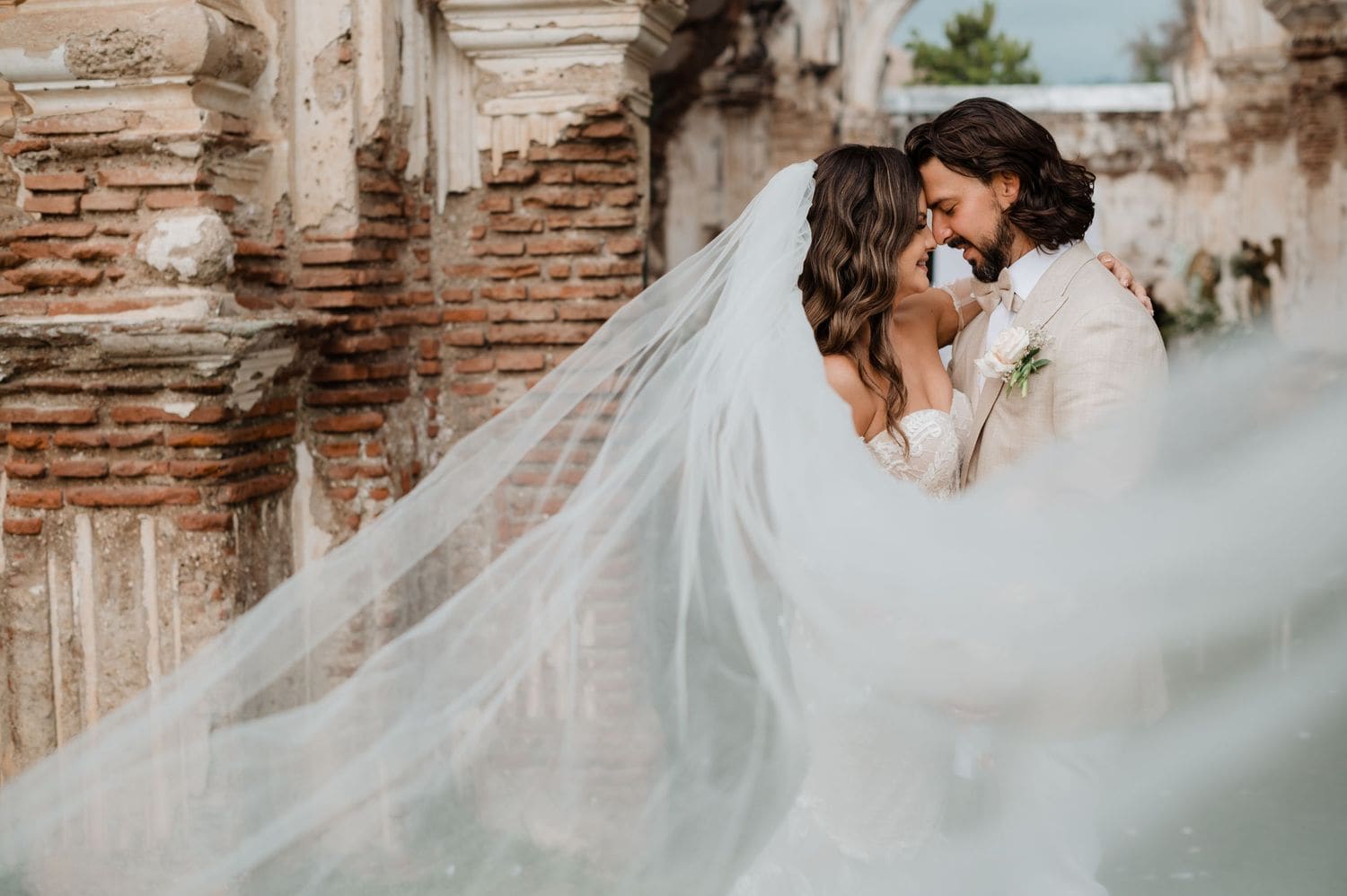 Bride and groom rest forehead-to-forehead as the long veil drapes across the lace gown in front of ruin walls.