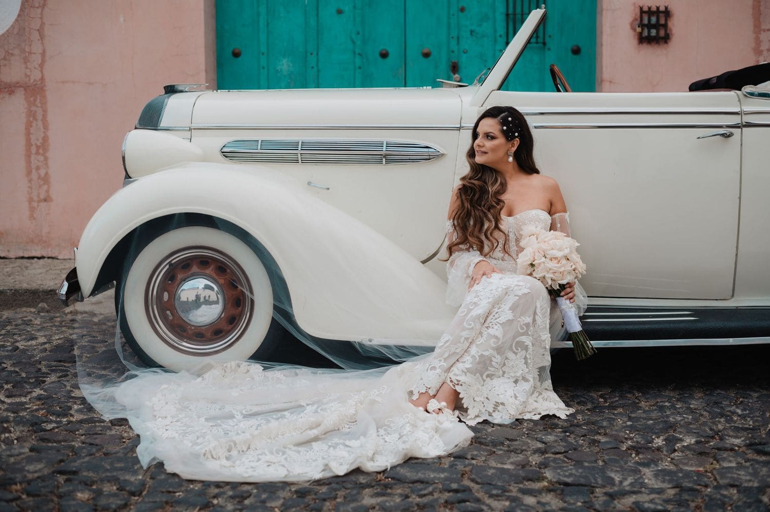 Bride alone seated against the side of a vintage white convertible in front of a turquoise colonial door.