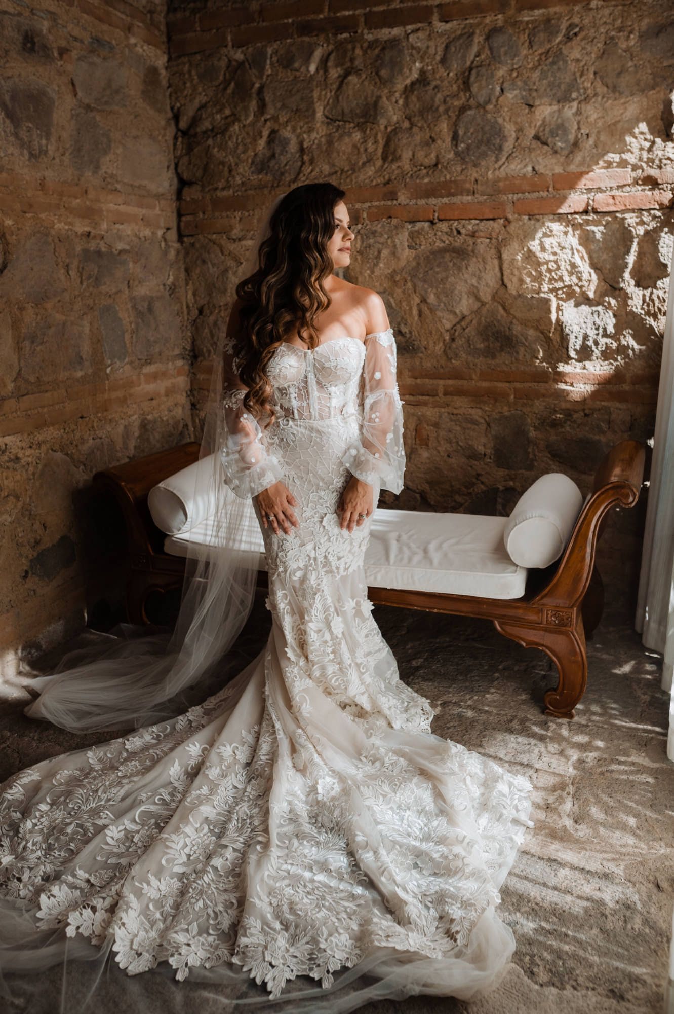 Bride in lace mermaid gown with long train poses near a stone-walled chaise in a sunlit bridal suite in Antigua Guatemala.