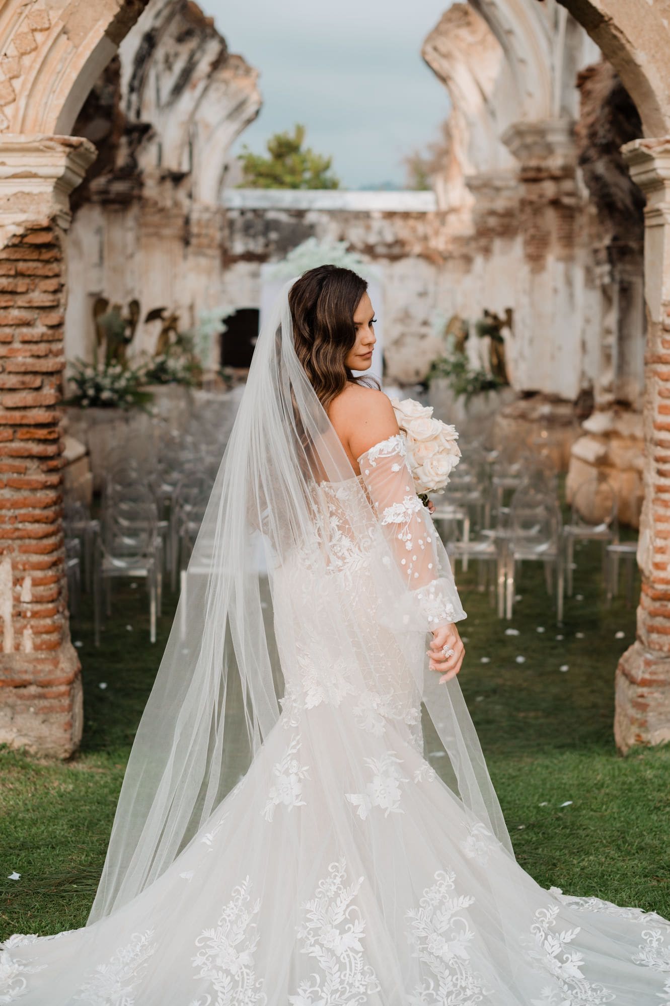 Bride from behind in lace mermaid gown with long veil drifting between brick ruin arches at Casa de la Ruina.