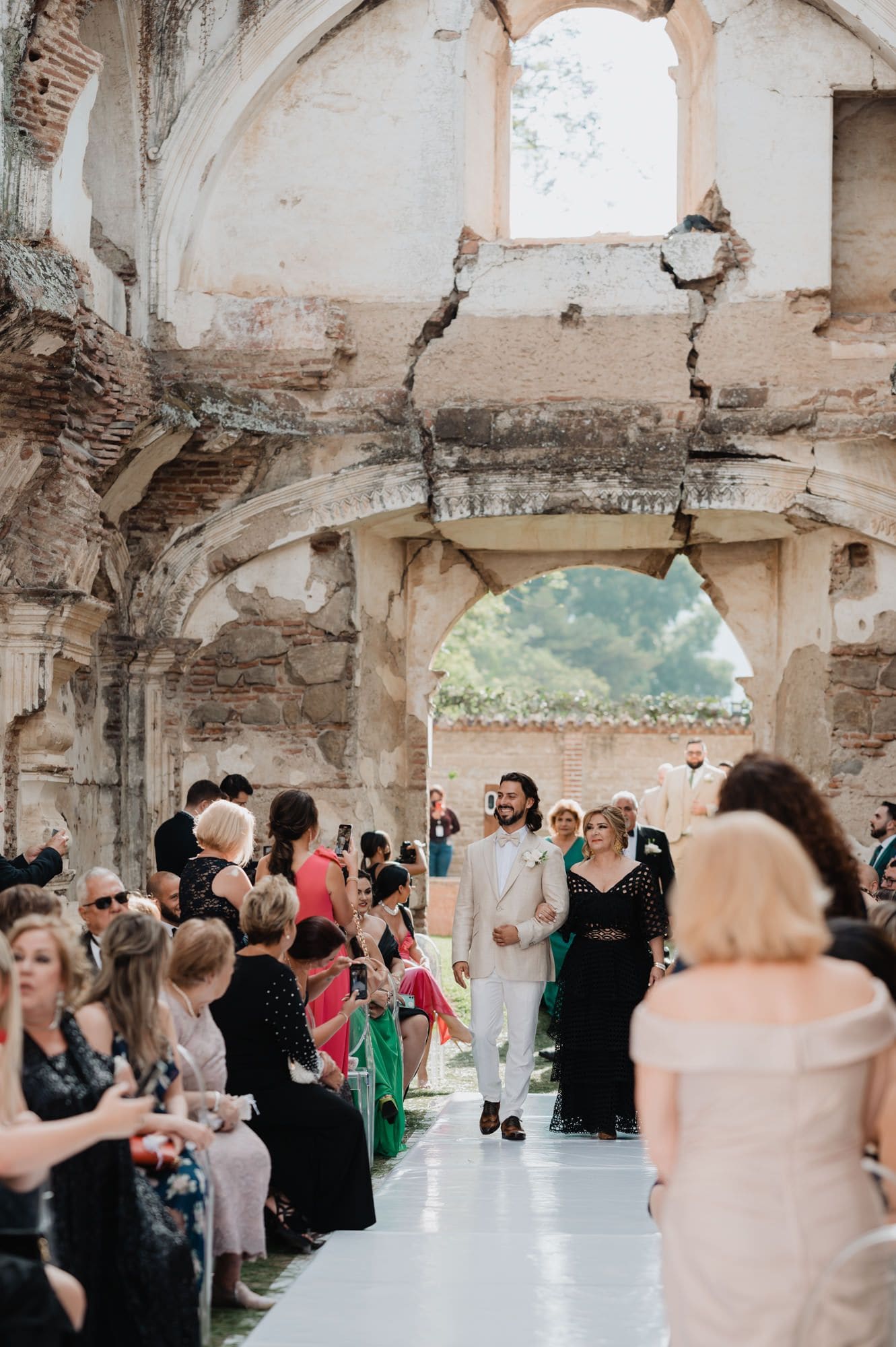 Groom in beige suit walks down the aisle with his mother under a cracked colonial arch at Casa de la Ruina.