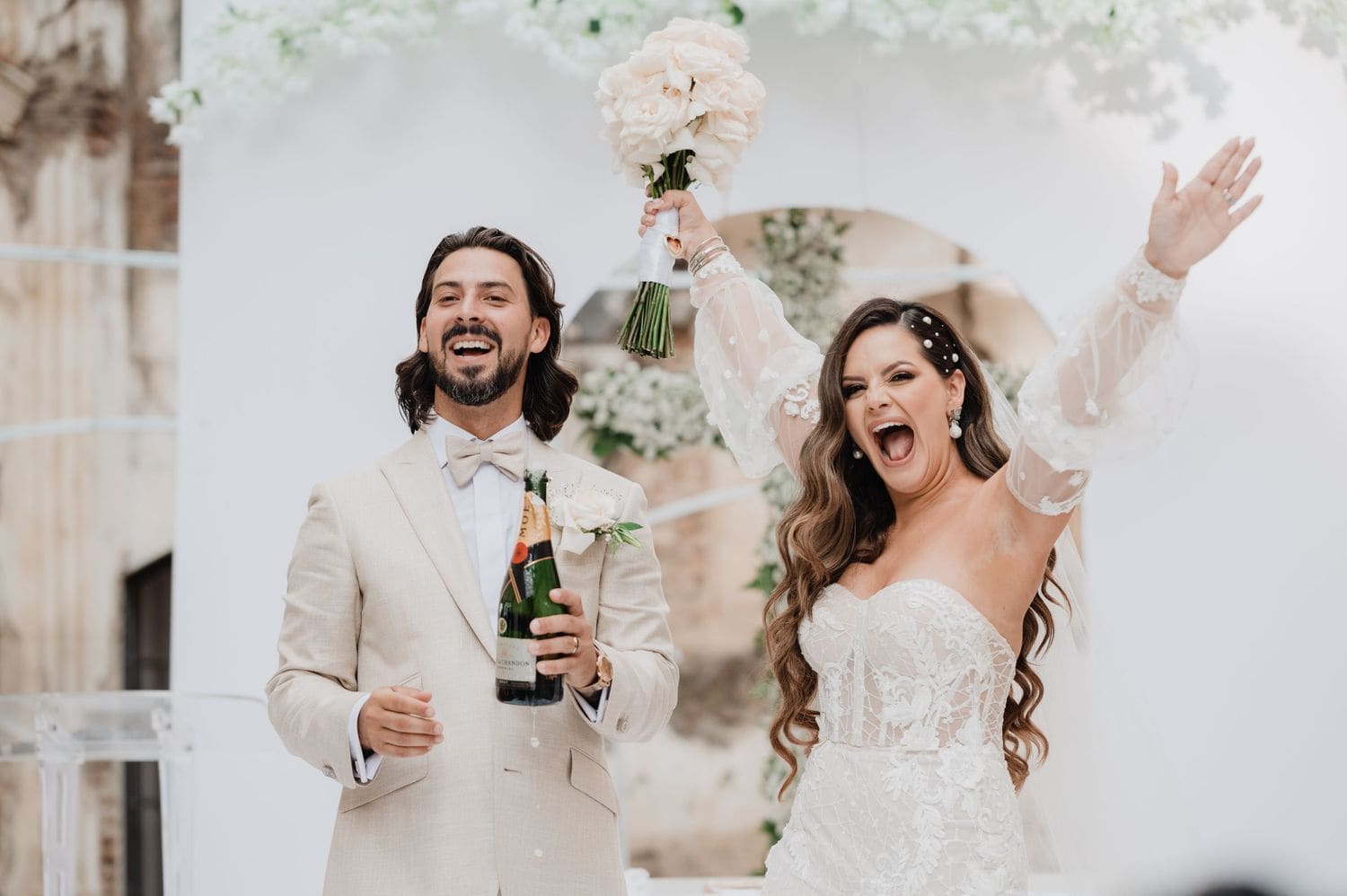 Couple laughs with arms raised and a bottle of champagne while celebrating the marriage at the altar.