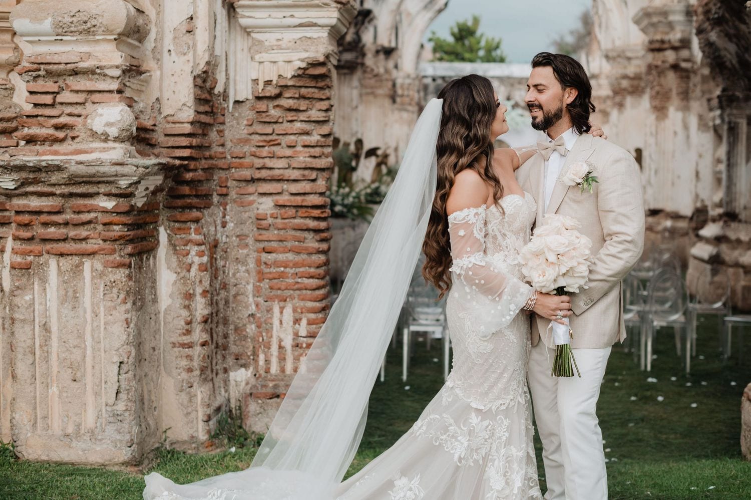Bride in lace gown with long veil shares a smiling embrace with her groom between brick ruin columns at Casa de la Ruina, Antigua Guatemala — editorial destination wedding portrait.