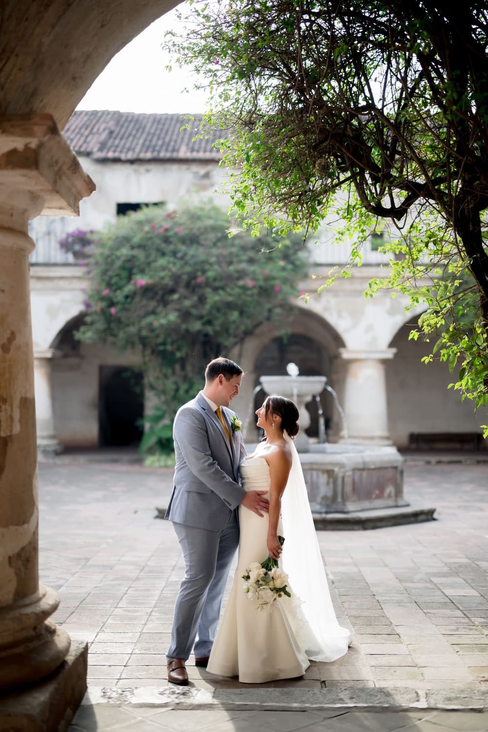 Bride in fitted strapless gown rests one hand on a moss-touched stone column, hora dorada light glowing on her face at Capuchinas Ruins.
