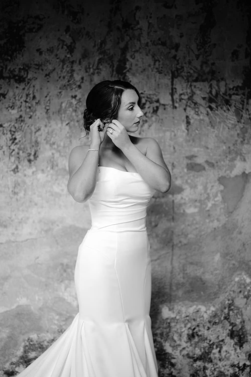 Bride in her white morning robe holds her bouquet, leaning gently against a stone wall while getting ready.