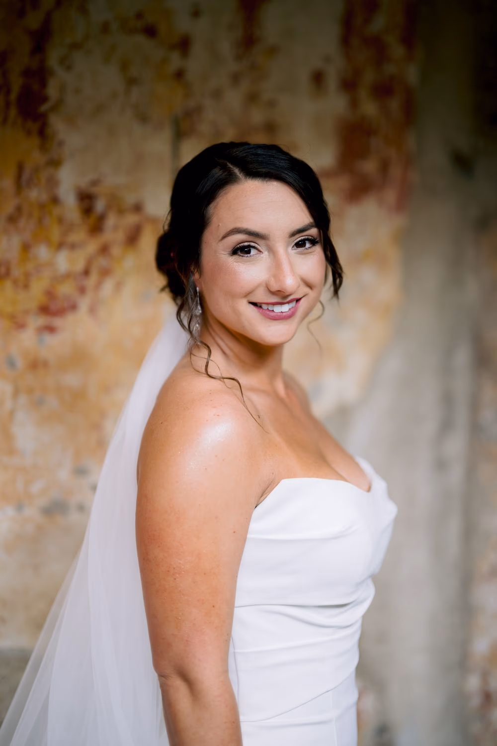 Bride lifts her veil with one hand, posed against an ochre patinated wall — full-length bridal portrait in La Antigua.