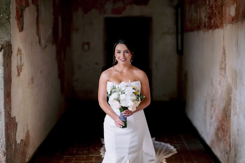 Bridal portrait — bride smiles softly against a patinated colonial wall, hair in a low bun and statement earrings catching the light.