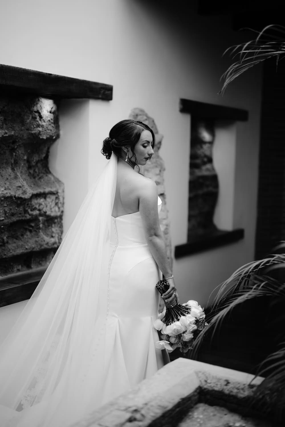 Bride stands in a long colonial corridor holding her white bouquet, the warm patina of the walls glowing around her.