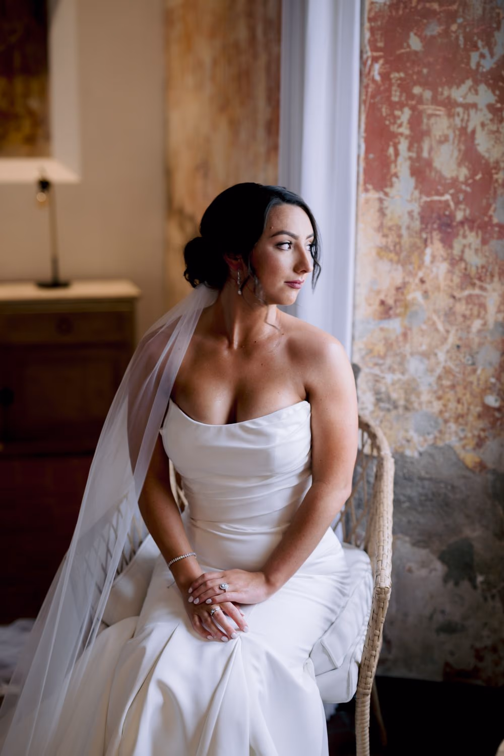 Black-and-white bridal portrait from behind — long cathedral veil cascading down the bride's back as she holds her bouquet by a stone window.