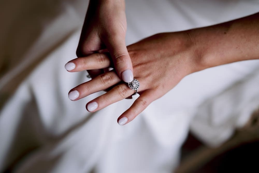Seated bride looks out a window in soft natural light, her wedding gown spilling across a woven chair — quiet pre-ceremony portrait.