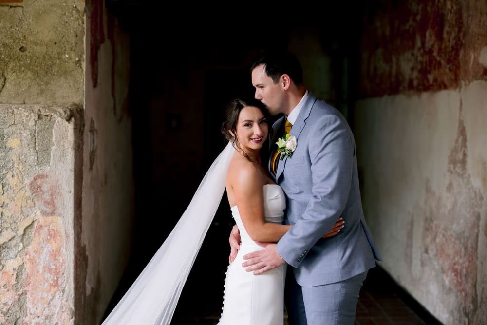 First look — bride and groom hold hands around a colonial pillar, both grinning before they see each other for the first time.