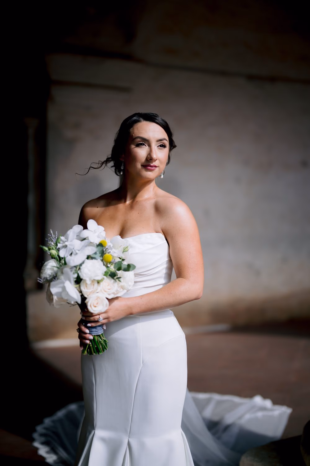 Bride pauses between stone columns of the convent cloister, profile lit by the afternoon glow, holding her white bouquet.