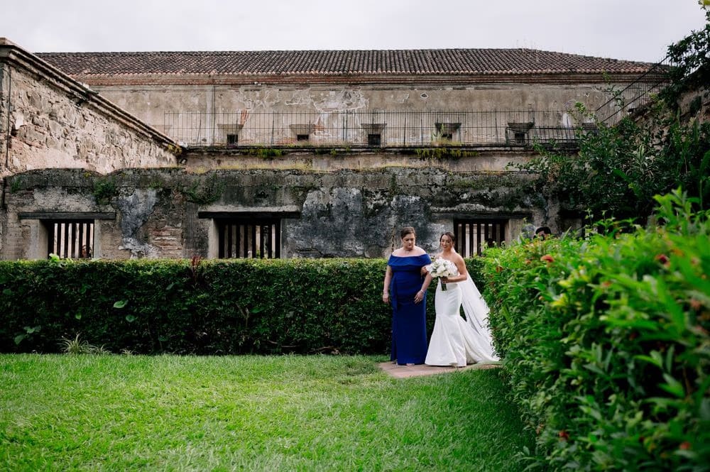 Bride and groom face the officiant beneath a tall floral arch as guests look on during the ceremony at Capuchinas Ruins.