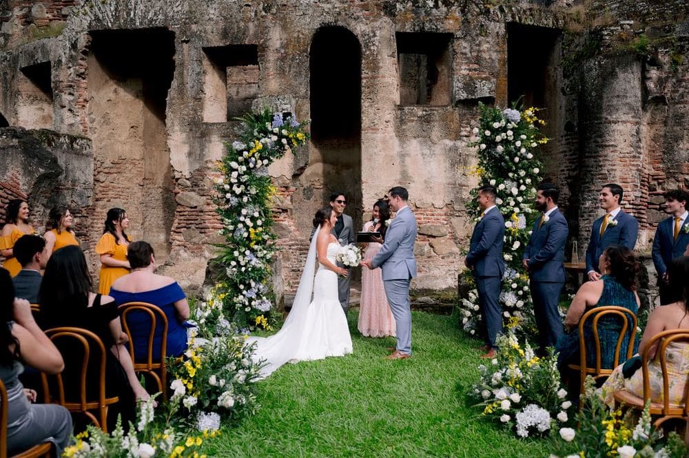 Bride walks toward the ceremony beside her mother, the convent ruins rising behind a green hedge.