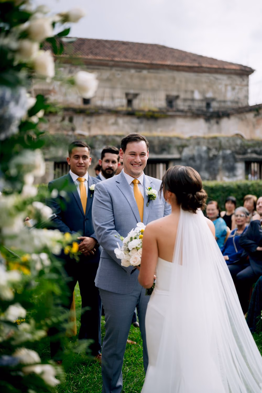 Wedding ceremony in progress — bride and groom hold hands at the altar framed by lush floral arches, bridal party flanking each side.