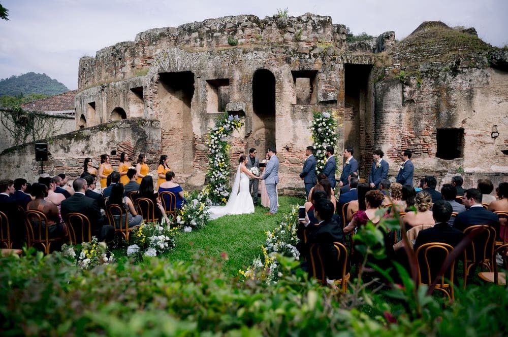 Wide ceremony shot — guests turn to watch the bride and groom exchange vows as the ruined walls and volcano rise behind them.