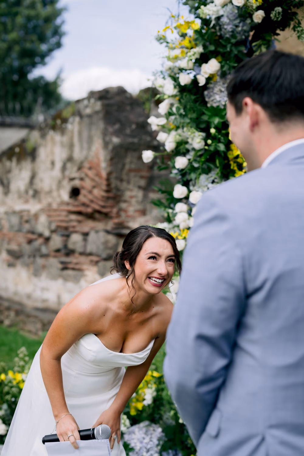 Bride leans forward laughing through her vows, microphone in hand, the groom listening with a wide smile.