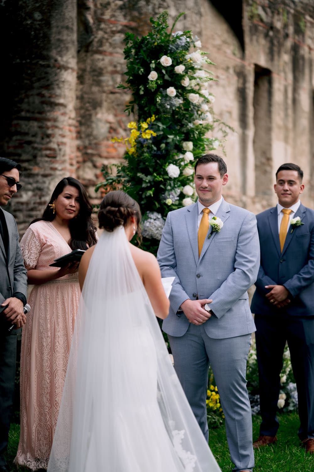 Groom listens to the bride's vows with a soft smile as the officiant stands beside them at the floral altar.