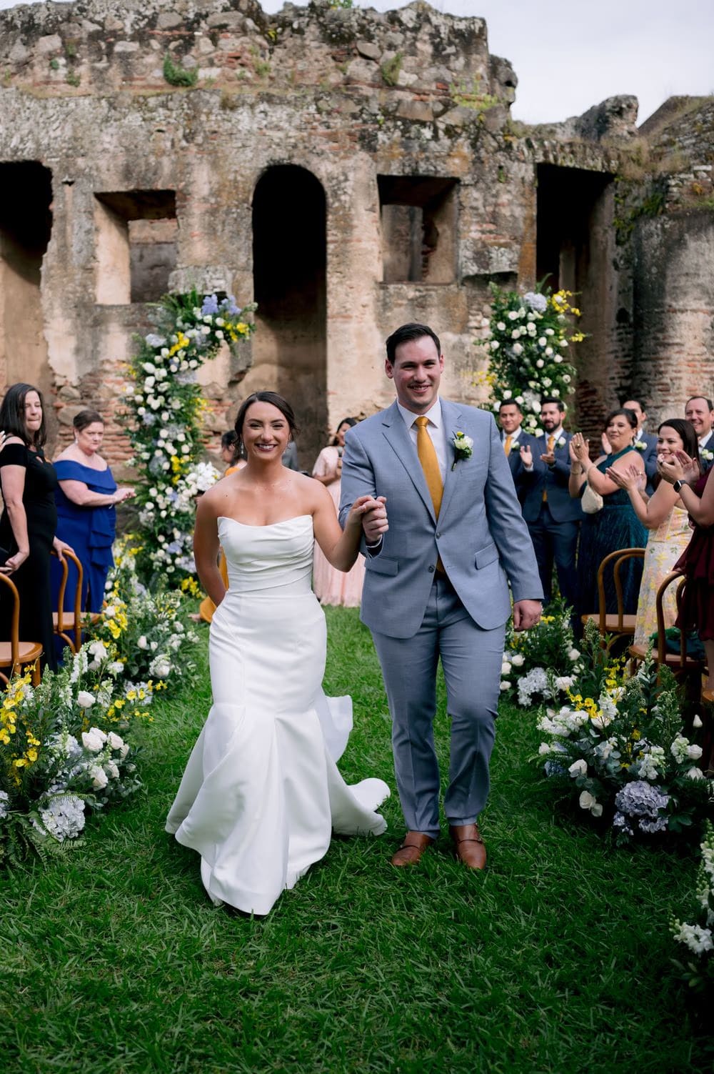 Newlyweds walk down the aisle hand in hand, beaming as guests applaud beneath the ruined arches.