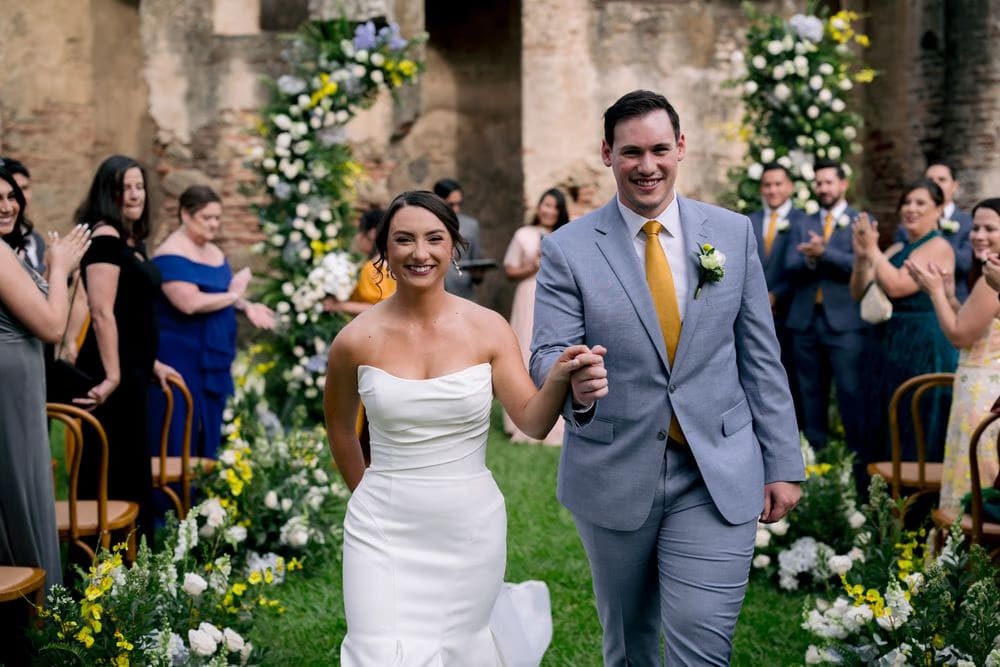 Close-up of the recessional — bride and groom laughing as they walk down the aisle through clapping guests.