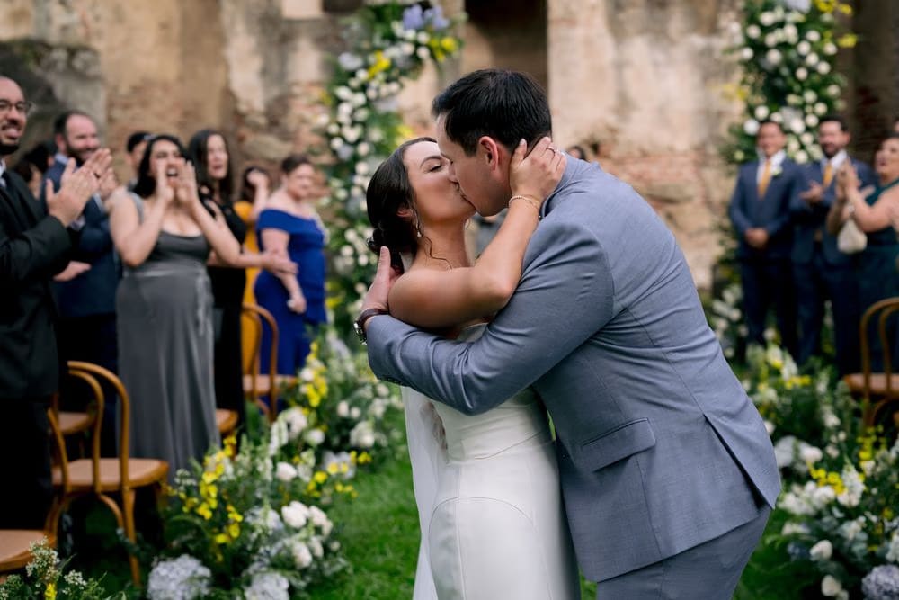 First kiss as a married couple — bride and groom share a deep kiss surrounded by cheering guests at Capuchinas Ruins.