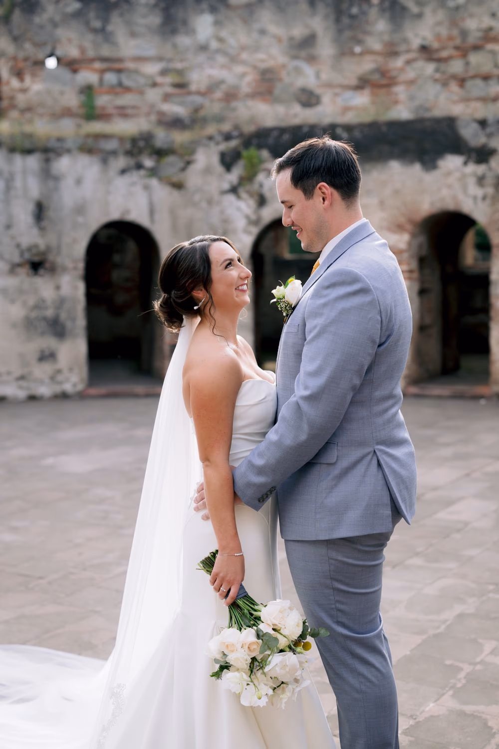 Bride and groom look at each other softly in a stone courtyard under the afternoon sun — quiet portrait moment after the ceremony.